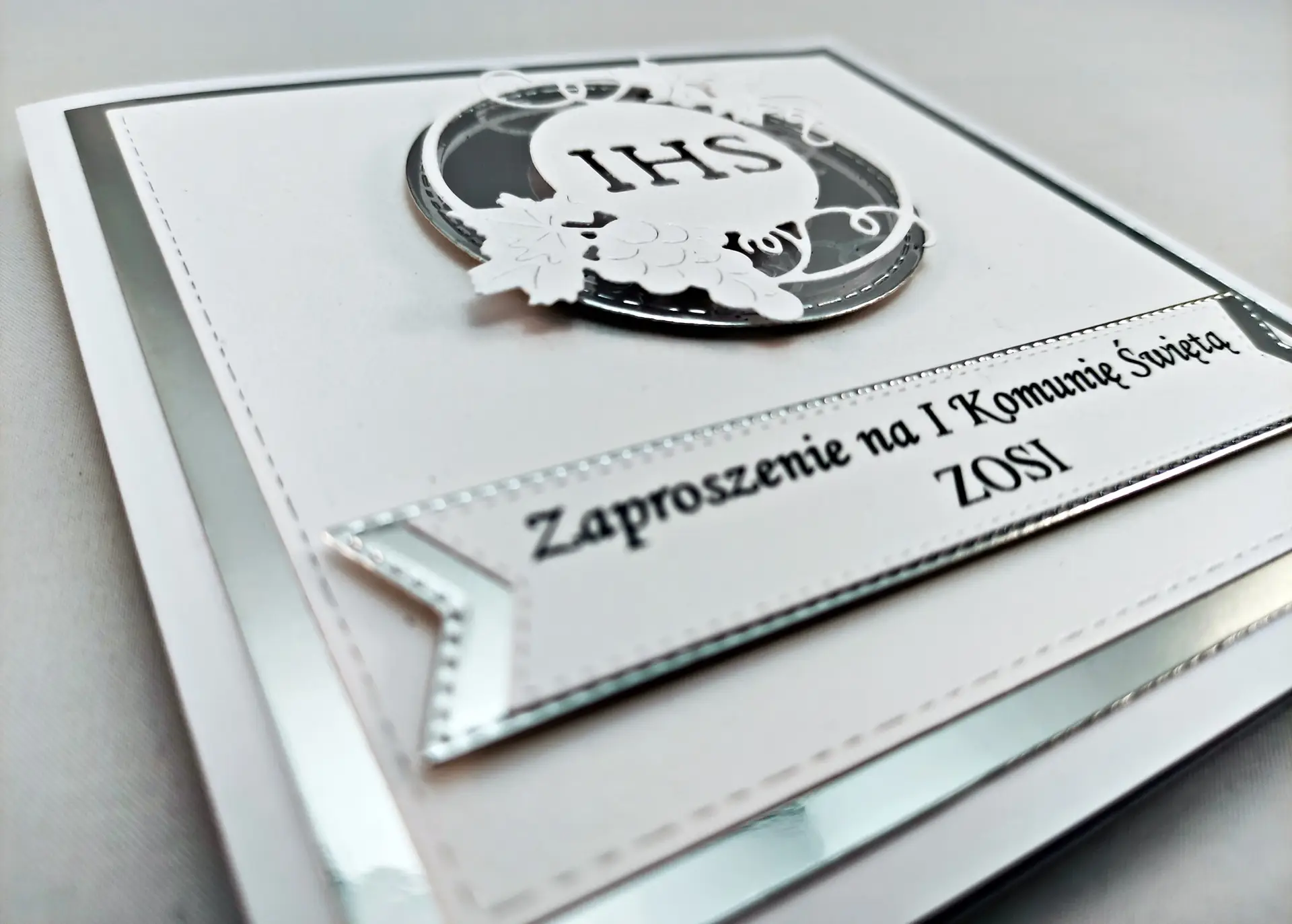 Zbliżenie zaproszenia na Pierwszą Komunię Świętą z symbolem IHS w srebrnym okręgu, na perłowym białym papierze z lustrzanym srebrnym tłem, delikatne i eleganckie wykończenie.