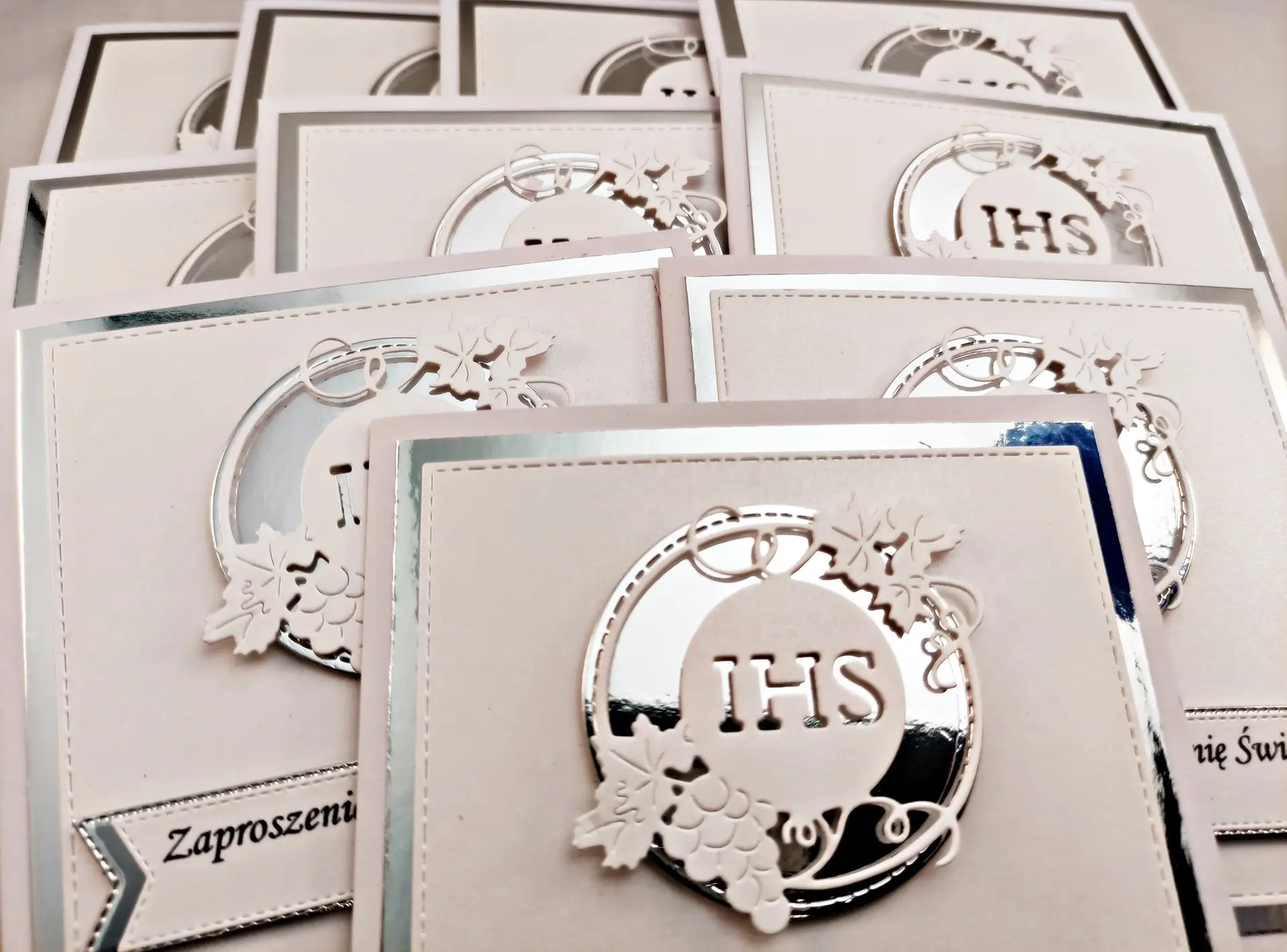 Kilka srebrnych zaproszeń komunijnych z motywem IHS w okręgu, ułożonych w rzędzie, widok z góry, połączenie perłowej bieli i lustrzanego srebra.