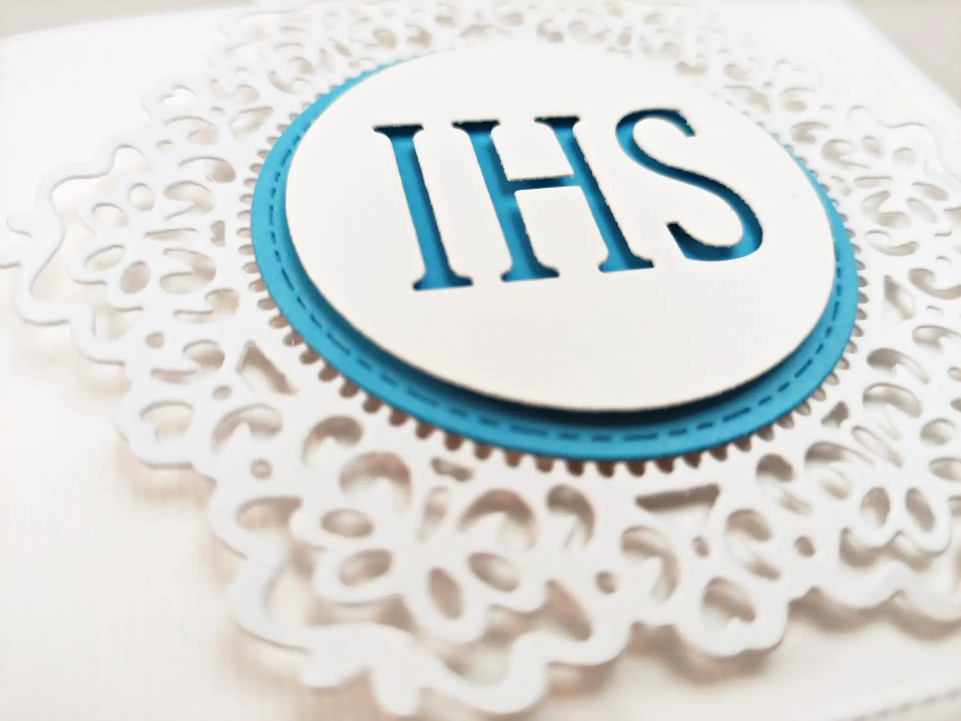 Zbliżenie niebieskiego zaproszenia komunijnego, pokazujące biały koronkowy ornament i centralnie umieszczony symbol IHS, widoczne detale wycinania.