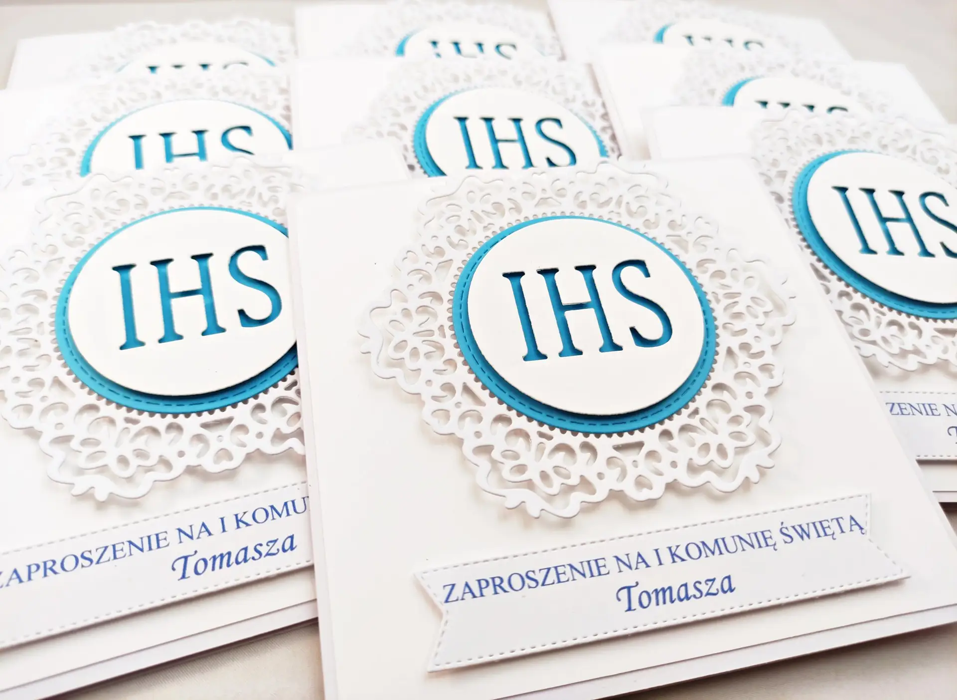 Kilka niebieskich zaproszeń na Pierwszą Komunię Świętą z koronkowym ornamentem i IHS, ułożonych obok siebie, widok z góry, spójna seria eleganckich zaproszeń.