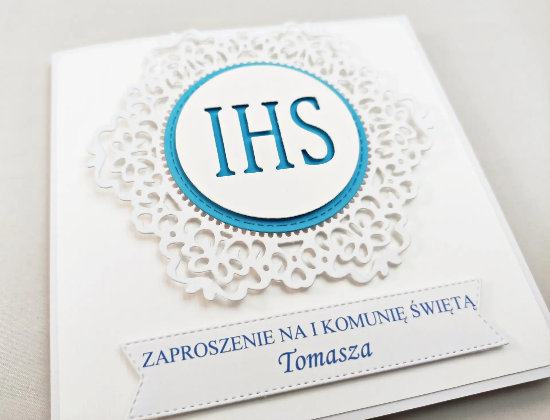 Niebieskie zaproszenie komunijne z białą koronką i symbolem IHS, sfotografowane z przodu, elegancki wzór z dekoracyjnym ornamentem na tle intensywnego koloru.