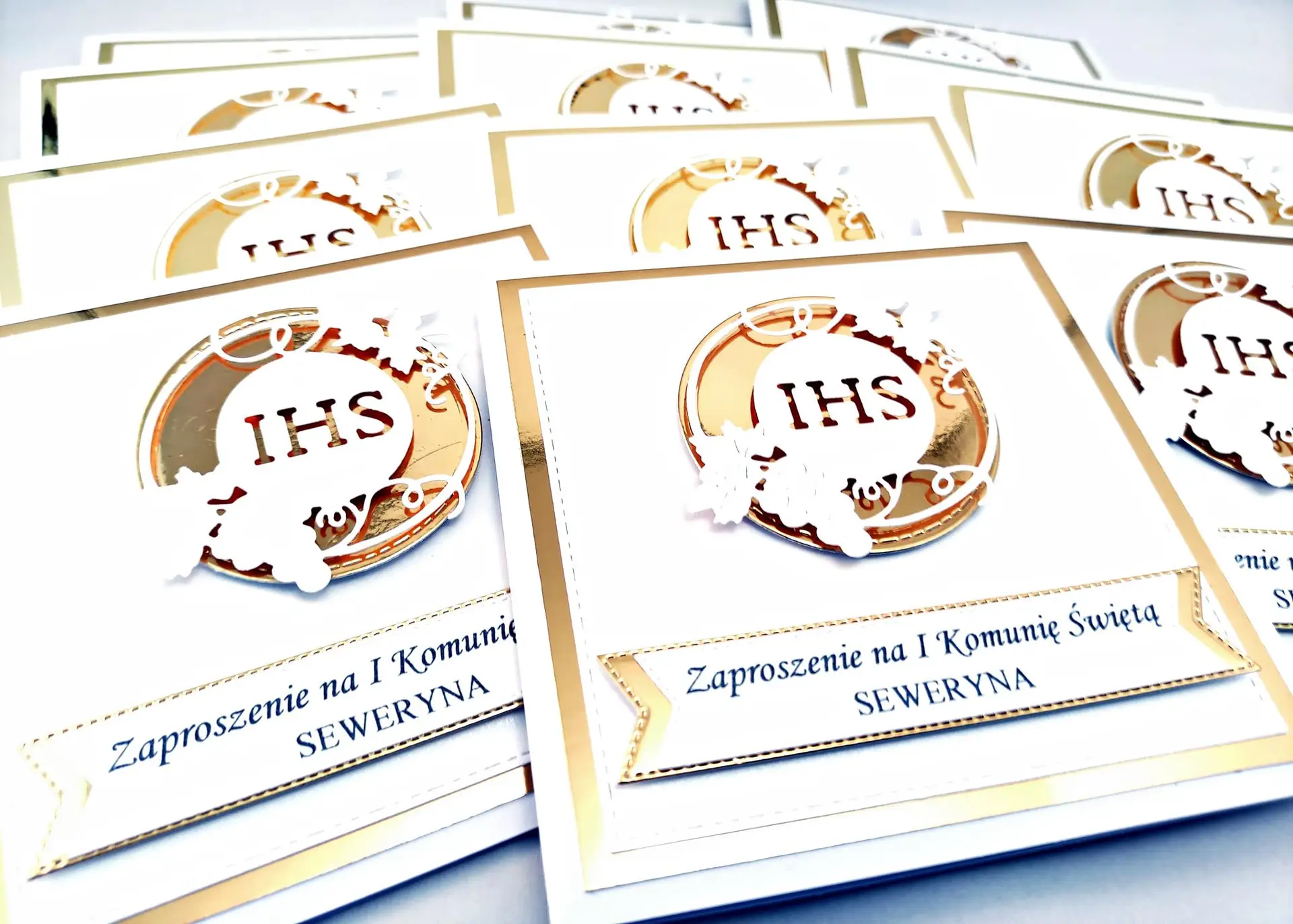 Kilka złotych zaproszeń komunijnych z motywem IHS w okręgu, ułożonych obok siebie na jasnym tle, widok z góry, perłowa biel połączona z lustrzanym złotem.