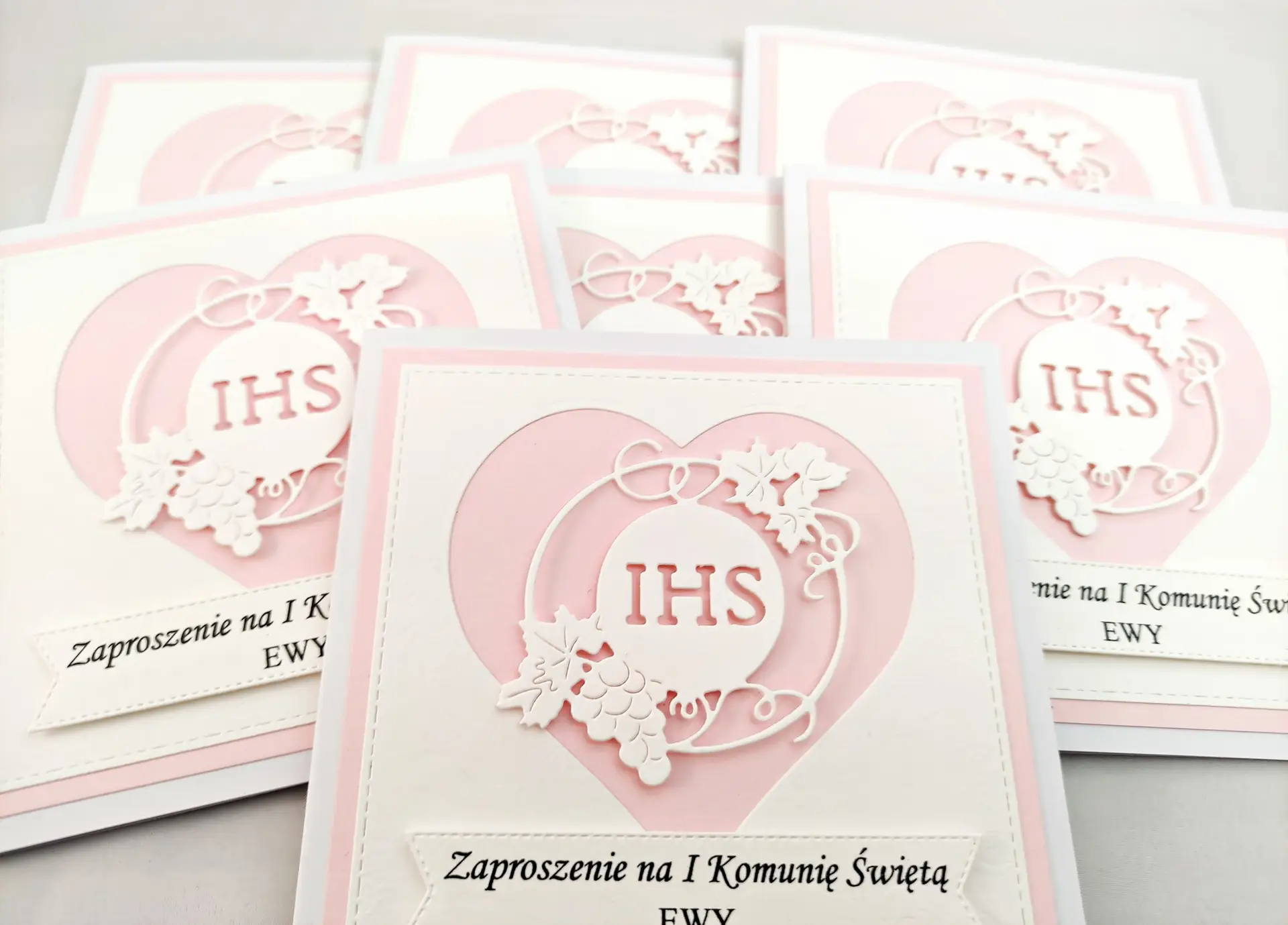 Kilka różowych zaproszeń na Pierwszą Komunię Świętą z sercem i symbolem IHS, ułożonych obok siebie, widok z góry.
