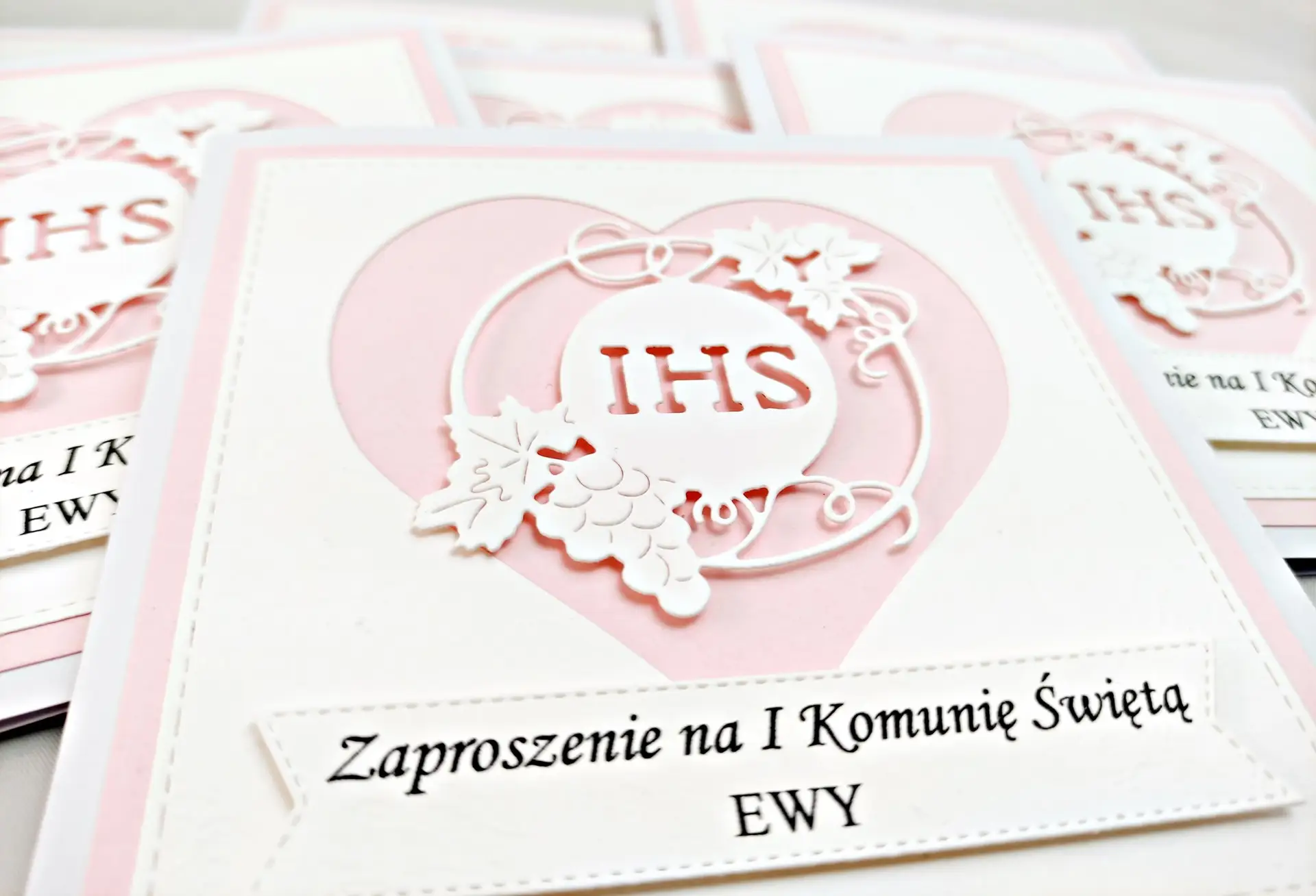 Zbliżenie różowego zaproszenia komunijnego dla dziewczynki z sercem i symbolem IHS na białym tle, delikatne dekoracyjne wykończenia.