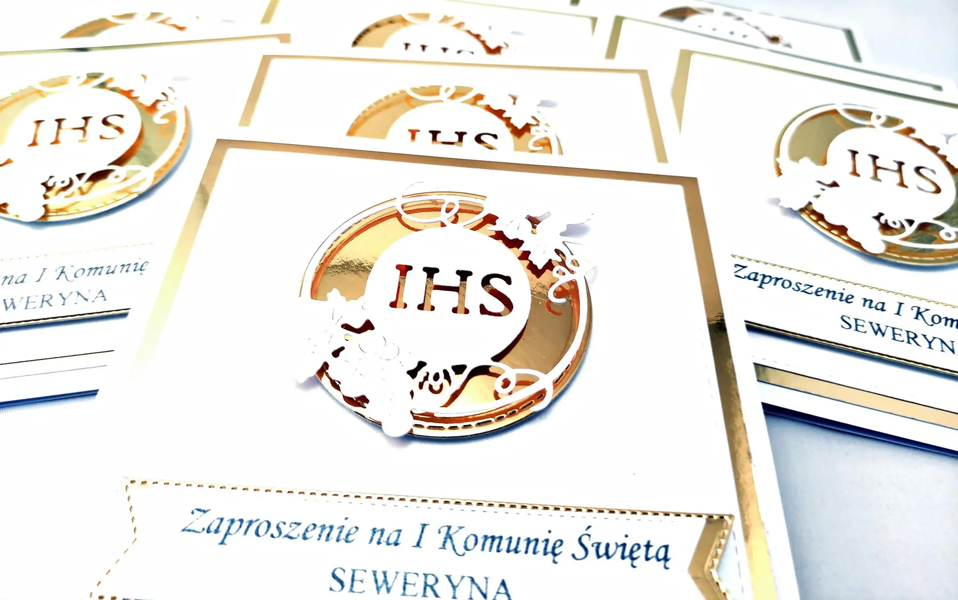 Zbliżenie zaproszenia na Pierwszą Komunię Świętą z motywem IHS w złotym okręgu, na perłowej bieli i lustrzanym złotym tle, eleganckie detale widoczne z bliska.