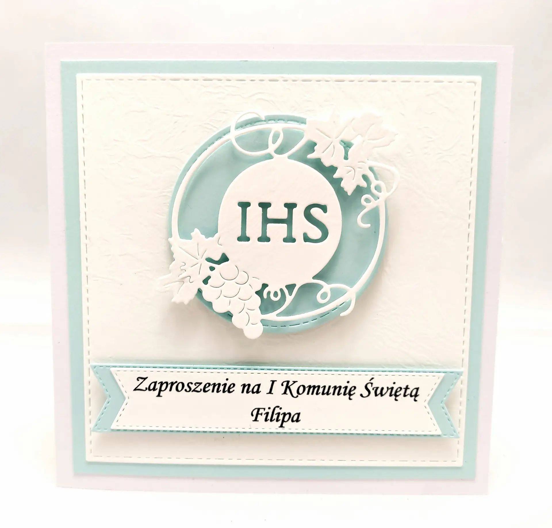 Błękitne zaproszenie na Pierwszą Komunię Świętą dla chłopca z motywem IHS w okręgu, wykonane z białego papieru wizytówkowego, eleganckie zaproszenie komunijne.