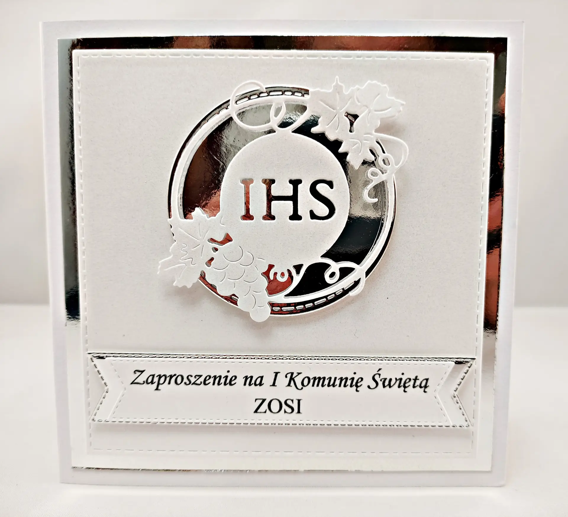 Perłowe zaproszenie komunijne w bieli z motywem IHS w srebrnym okręgu na lustrzanym tle, delikatne i eleganckie zaproszenie na Pierwszą Komunię Świętą.