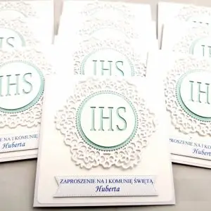 Kilka jasnobłękitnych zaproszeń na Pierwszą Komunię Świętą z białym koronkowym ornamentem i symbolem IHS, ułożonych w rzędzie, widok z góry, delikatna morska kolorystyka.