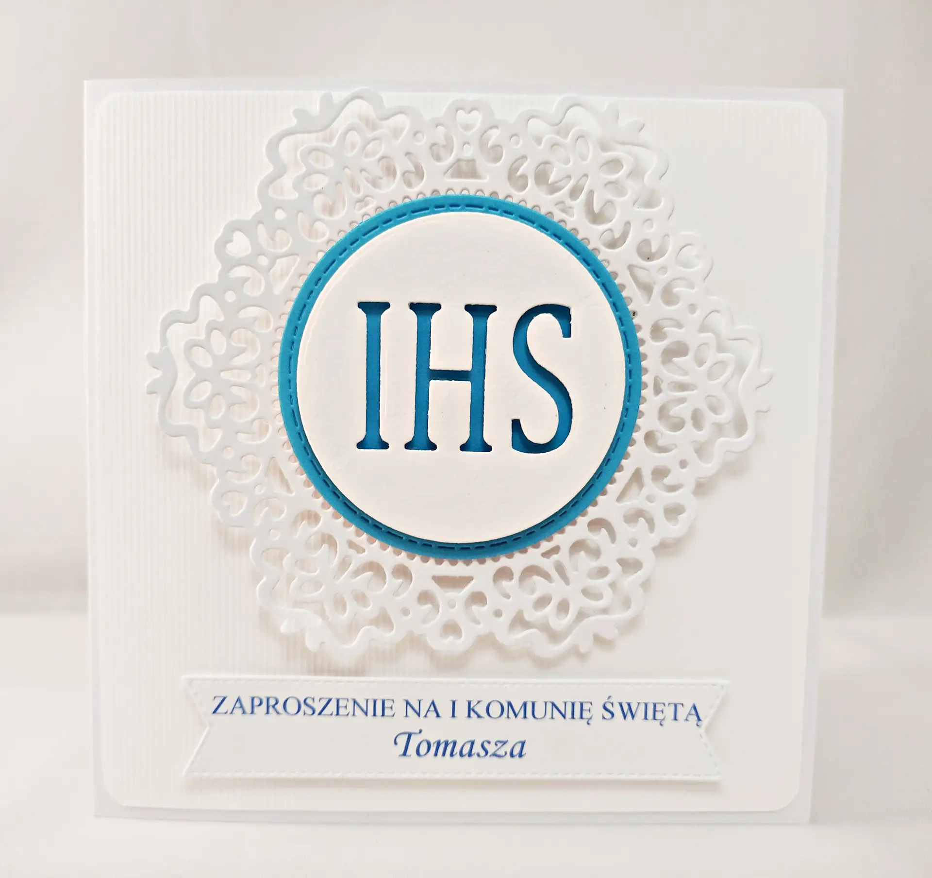Niebieskie zaproszenie na Pierwszą Komunię Świętą z białym koronkowym ornamentem i symbolem IHS, kwadratowe, eleganckie zaproszenie na papierze wizytówkowym.
