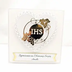 Biało-złote zaproszenie na Pierwszą Komunię Świętą z ażurowym sercem i symbolem IHS w otoczeniu winorośli, pojedyncze zaproszenie sfotografowane od frontu.