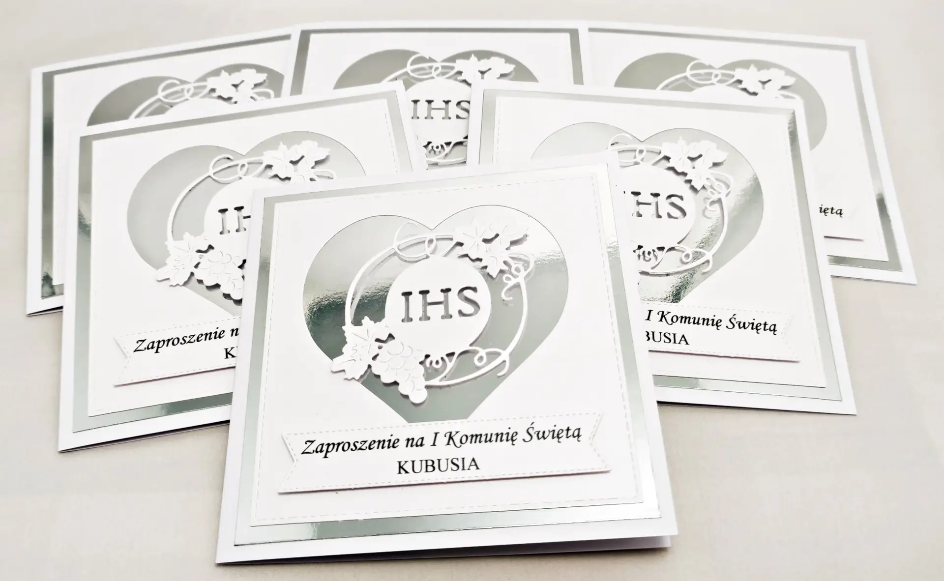 Zaproszenie na Pierwszą Komunię Świętą w kolorze perłowej bieli, z motywem serca i symbolem IHS w lustrzanym srebrze, eleganckie i delikatne zaproszenie komunijne.