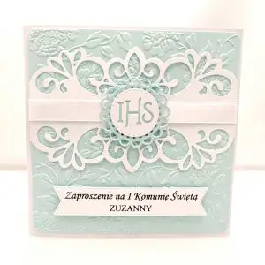 Błękitne zaproszenie na Pierwszą Komunię Świętą dla chłopca, z tłoczonym tłem, dekoracyjnymi ornamentami i symbolem IHS umieszczonym na jasnej satynowej wstążce.