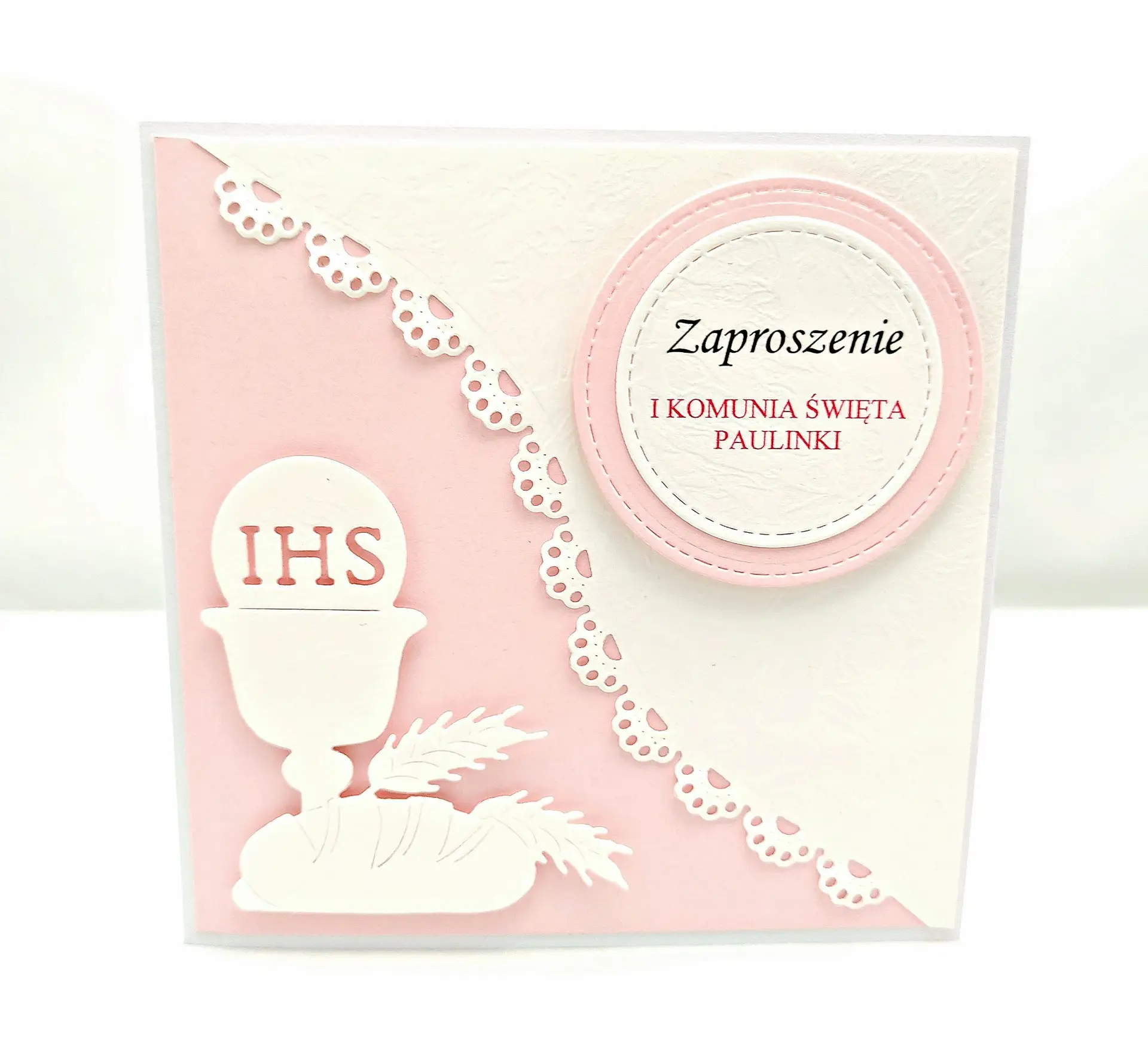 Różowe zaproszenie na Pierwszą Komunię Świętą dla dziewczynki, z motywem kielicha IHS, skośnym różowym tłem i ażurową koronką na białym papierze wizytówkowym.