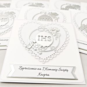 Kilka biało-srebrnych zaproszeń na Pierwszą Komunię Świętą z ażurowym sercem i symbolem IHS w otoczeniu winorośli, ułożonych obok siebie, widok z góry.