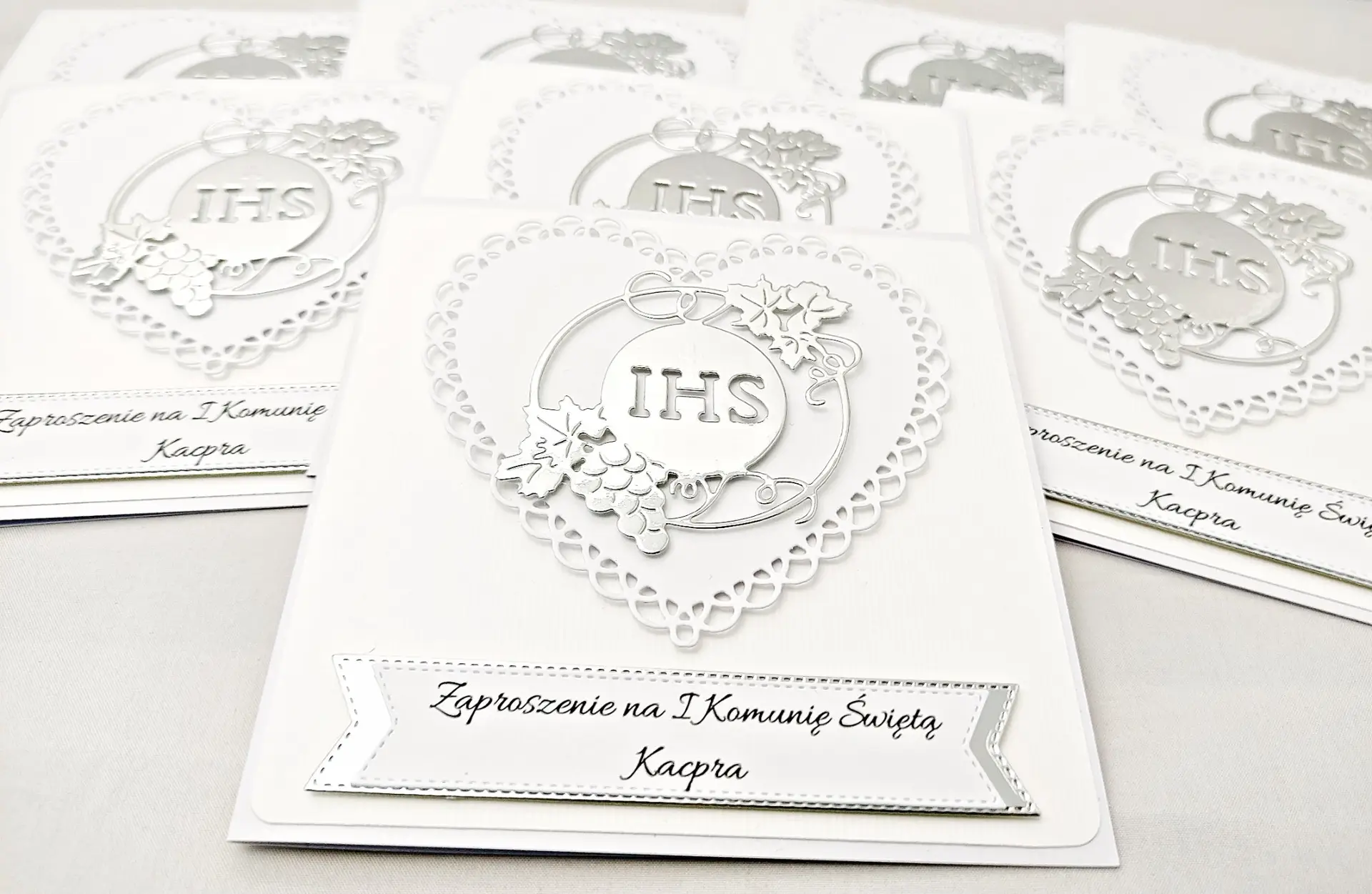 Kilka biało-srebrnych zaproszeń na Pierwszą Komunię Świętą z ażurowym sercem i symbolem IHS w otoczeniu winorośli, ułożonych obok siebie, widok z góry.