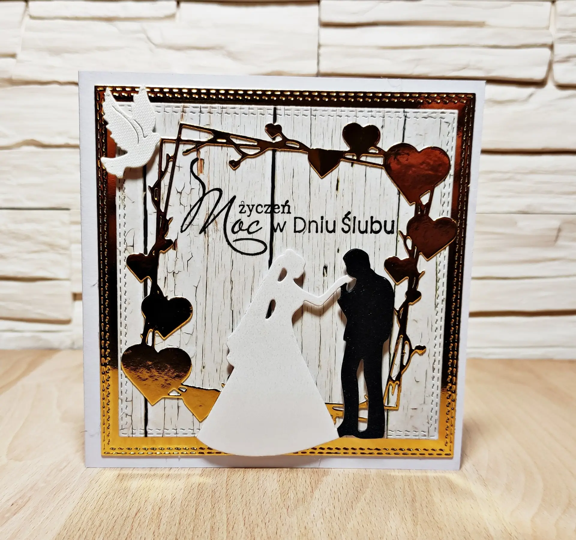 kwadratowa kartka ślubna ręcznie robiona z sylwetką pary młodej i girlandą wiszących serc, elegancka kartka na ślub 13,5x13,5 cm w złoto-białej kolorystyce