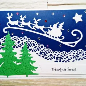 ażurowa kartka świąteczna z białymi saniami Mikołaja i reniferami na granatowym tle, z zielonymi choinkami i napisem Wesołych Świąt