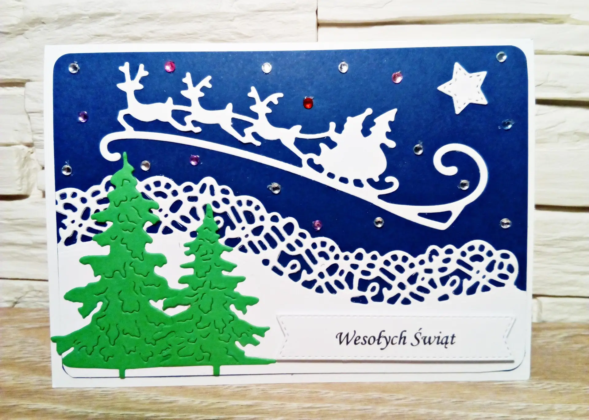 ażurowa kartka świąteczna z białymi saniami Mikołaja i reniferami na granatowym tle, z zielonymi choinkami i napisem Wesołych Świąt