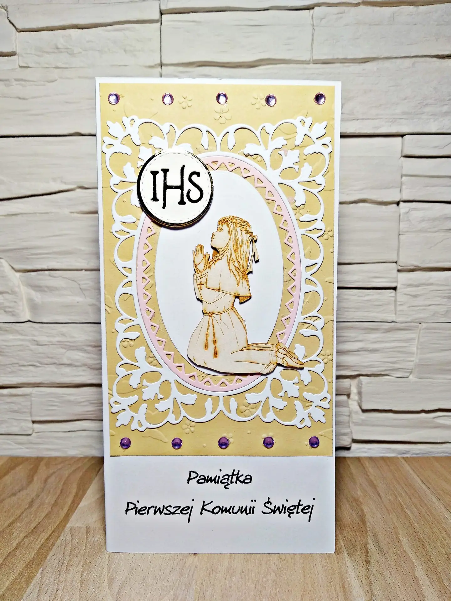 Ręcznie wykonana kartka komunijna DL z tekturową klęczącą dziewczynką w stroju komunijnym i napisem „Pamiątka Pierwszej Komunii Świętej”, subtelne komunijne zdobienia na dekoracyjnym tle
