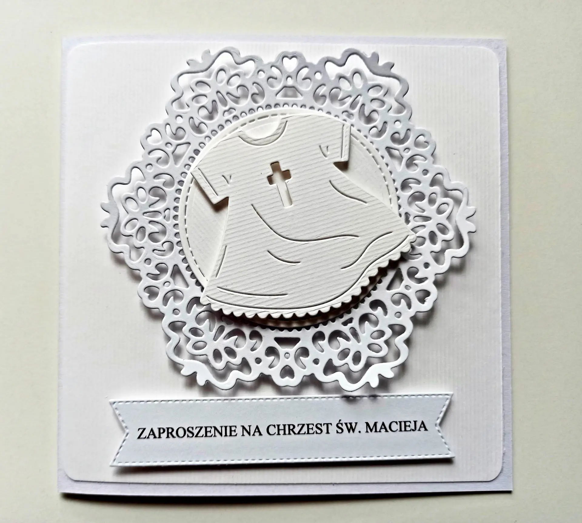 Uniwersalne zaproszenie na Chrzest Święty z motywem białej szatki do chrztu na tle koronki, całe w bieli, ręcznie robione zaproszenie