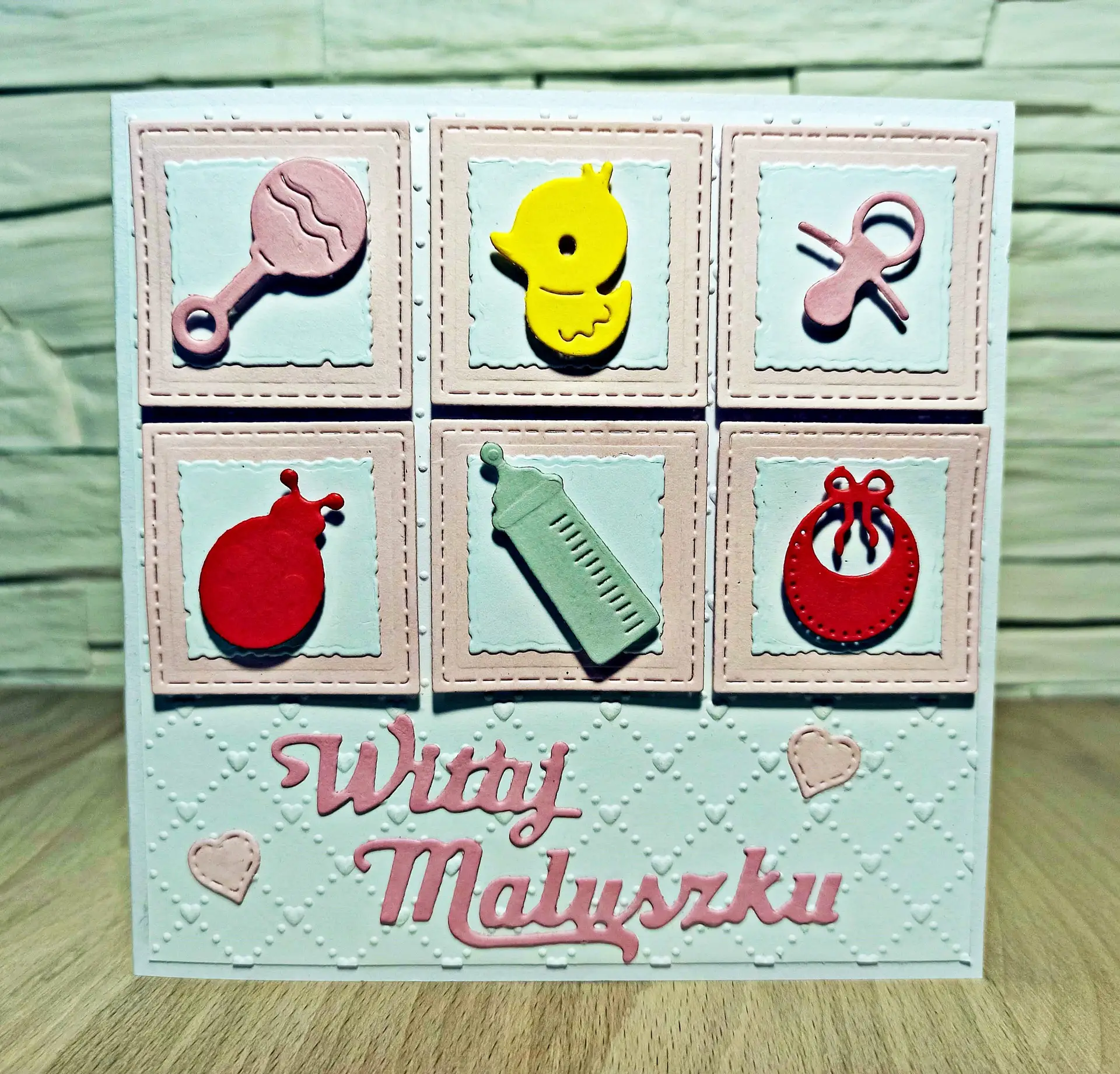 kwadratowa kartka gratulacyjna Witaj Maluszku z kolorowymi dziecięcymi gadżetami w małych kwadratach, ręcznie robiona kartka z okazji narodzin córki