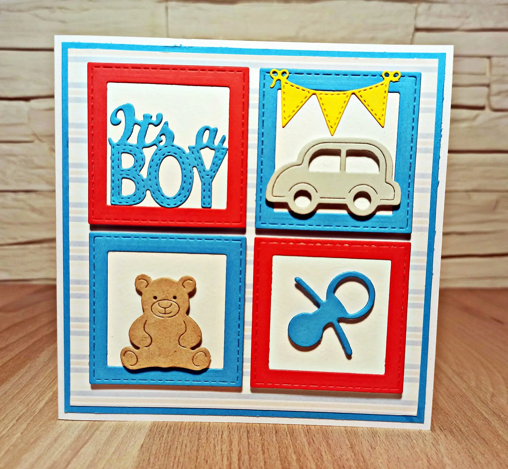 kwadratowa kartka gratulacyjna It's a Boy w niebiesko-czerwonej kolorystyce z misiem, samochodem i smoczkiem, ręcznie robiona kartka z okazji narodzin chłopca