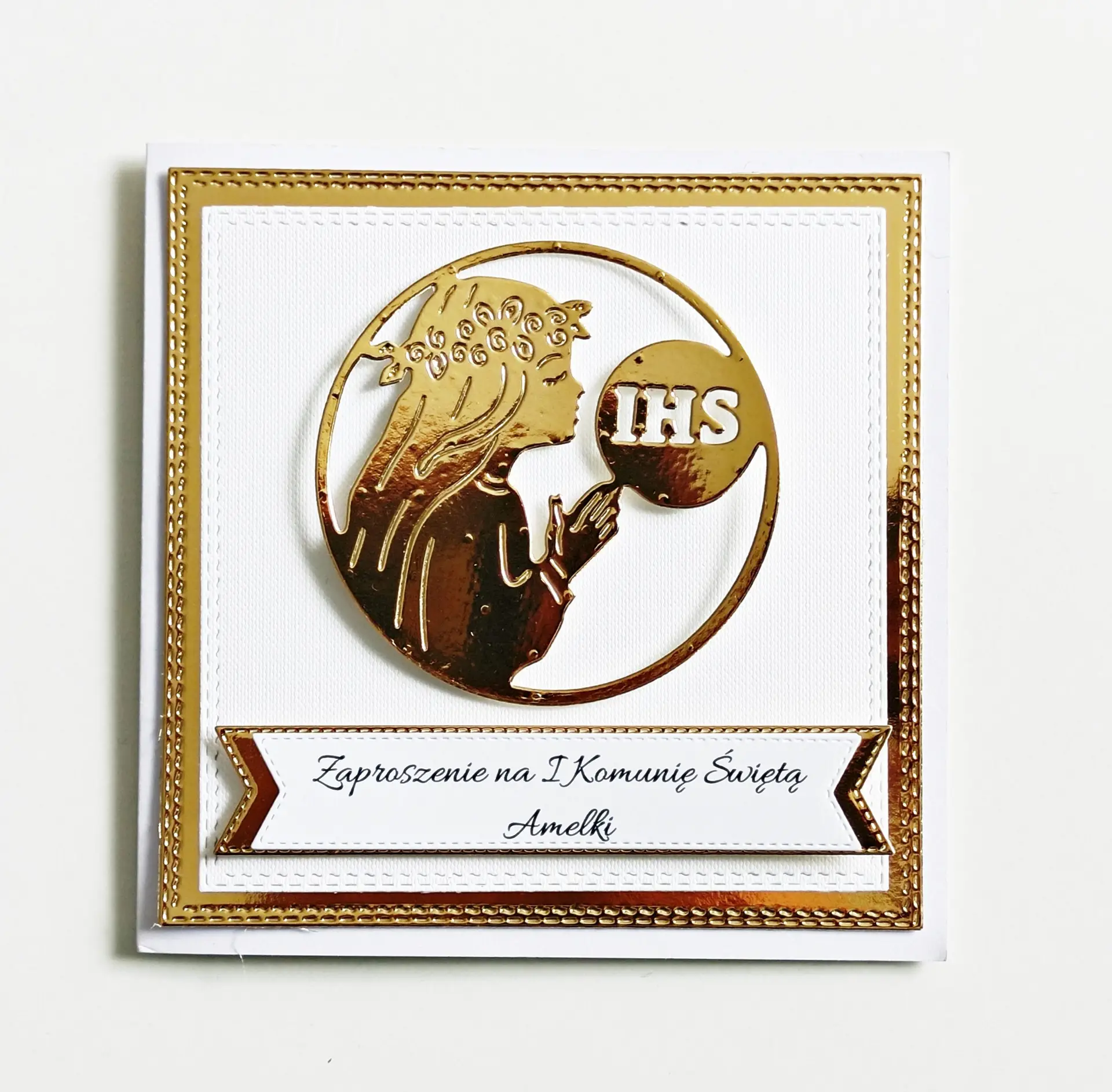 Zaproszenie na Pierwszą Komunię Świętą dla dziewczynki z sylwetką modlącej się dziewczynki i symbolem IHS, na białym papierze wizytówkowym z lustrzanym złotym tłem.
