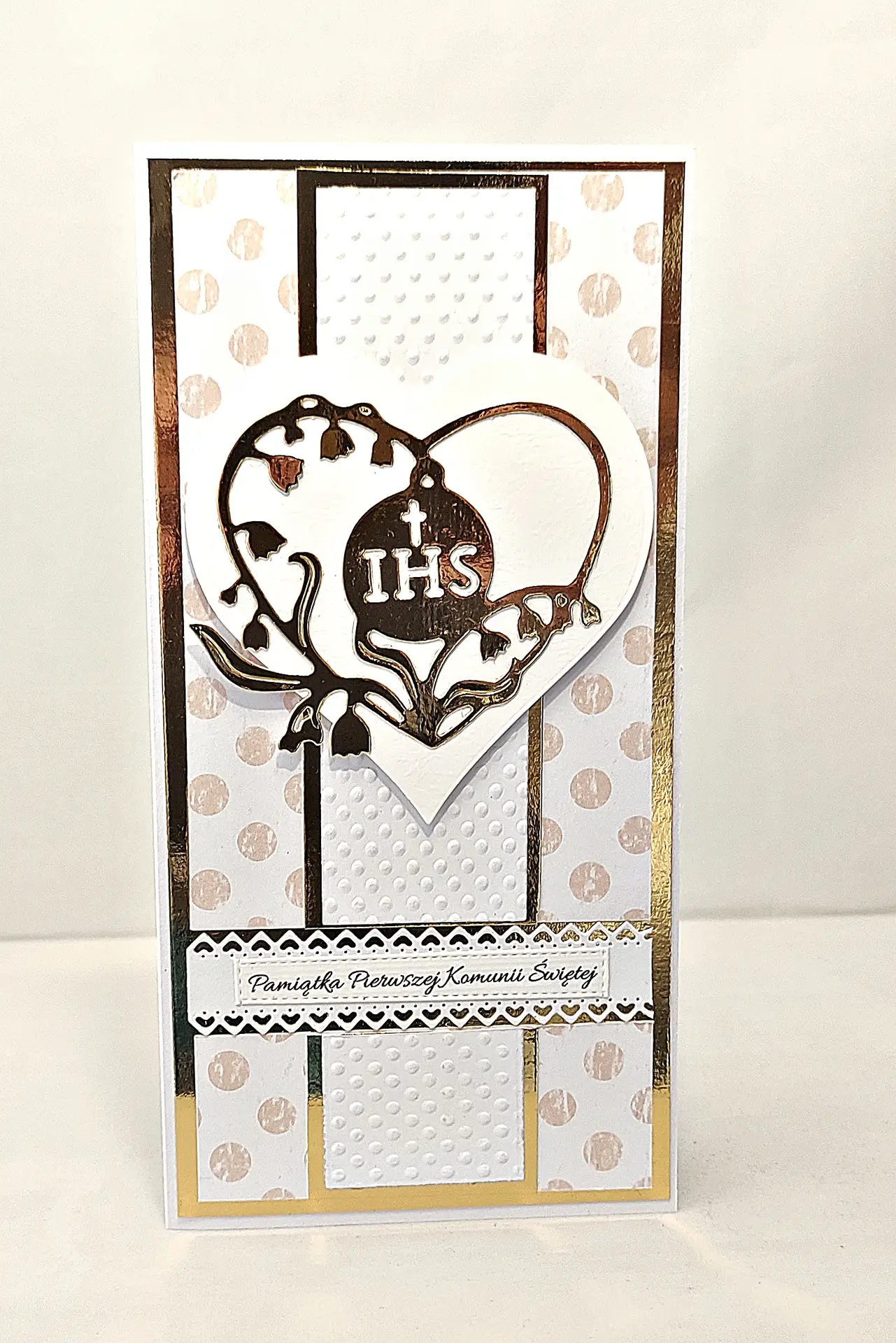 Ręcznie robiona kartka komunijna DL z motywem IHS w ozdobnym sercu i napisem „Pamiątka Pierwszej Komunii Świętej”, delikatne zdobienia i eleganckie tło