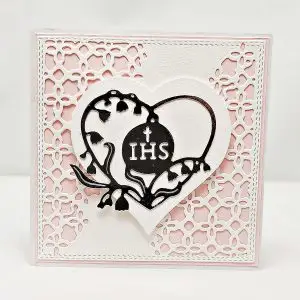 Ręcznie robiona kartka komunijna 13,5x13,5 dla dziewczynki, w pastelowym różu, z sercem i symbolem IHS na dekoracyjnym tle, delikatna pamiątka Pierwszej Komunii Świętej