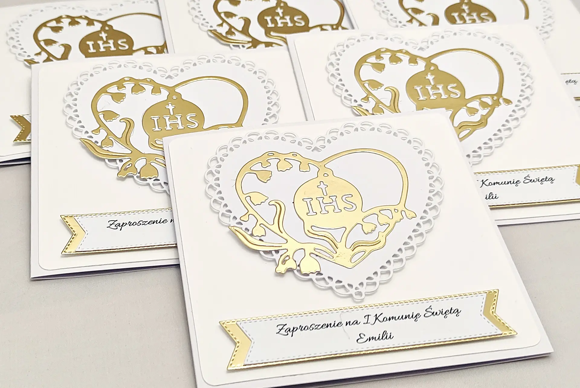 Kilka biało-złotych zaproszeń na Pierwszą Komunię Świętą z ażurowym sercem i sercem z konwalii z symbolem IHS, ułożonych obok siebie, widok z góry.