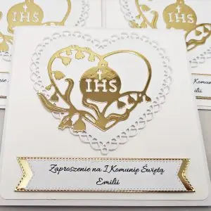 Zbliżenie biało-złotego zaproszenia na Pierwszą Komunię Świętą z ażurowym sercem i sercem z konwalii wokół symbolu IHS.