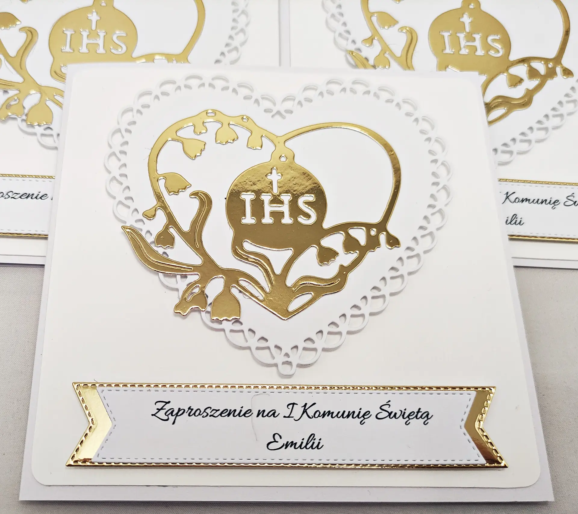Zbliżenie biało-złotego zaproszenia na Pierwszą Komunię Świętą z ażurowym sercem i sercem z konwalii wokół symbolu IHS.