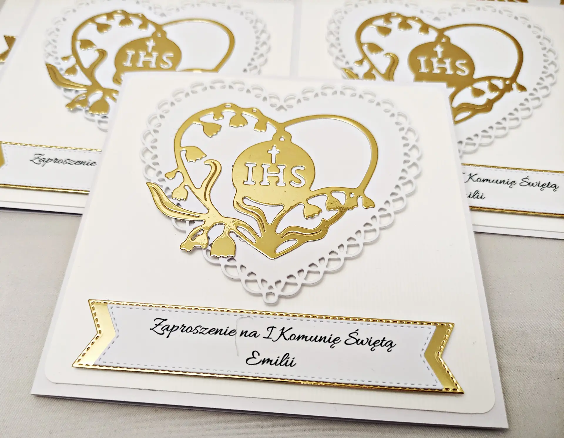 Zaproszenie komunijne w biało-złotej kolorystyce, pokazane z bliska, z ażurowym sercem i wnętrzem w formie serca z konwalii z symbolem IHS.