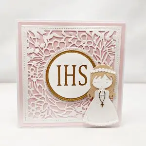 Delikatna kartka komunijna dla dziewczynki z motywem IHS i dziewczynką, ozdobiona ażurowym tłem i pastelowymi akcentami, elegancka pamiątka Pierwszej Komunii Świętej