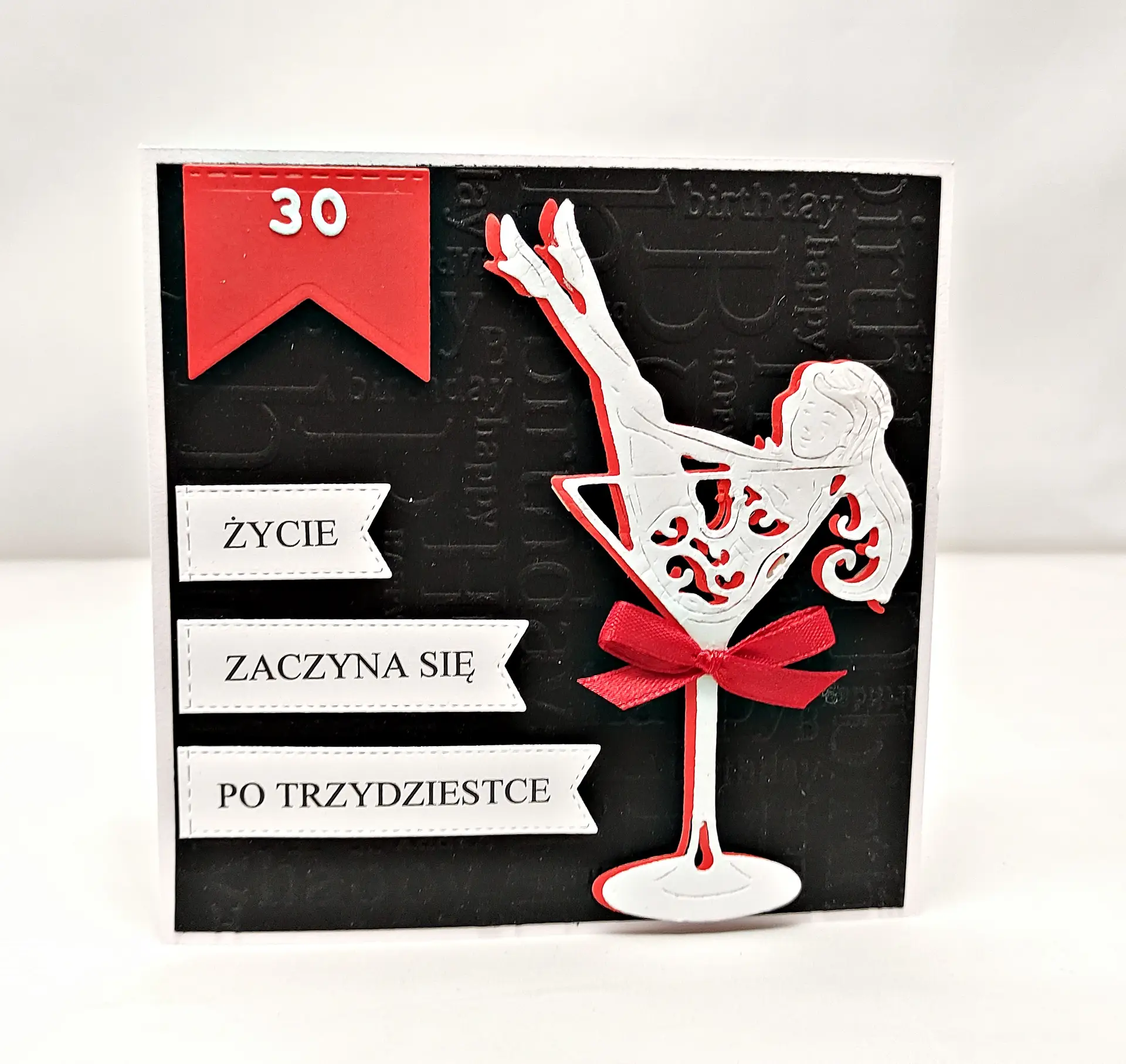 Ręcznie robiona kartka na 30 urodziny z napisem „Życie zaczyna się po trzydziestce”, czarne tłoczone tło, czerwono-biały kieliszek z sylwetką kobiety i czerwoną kokardką