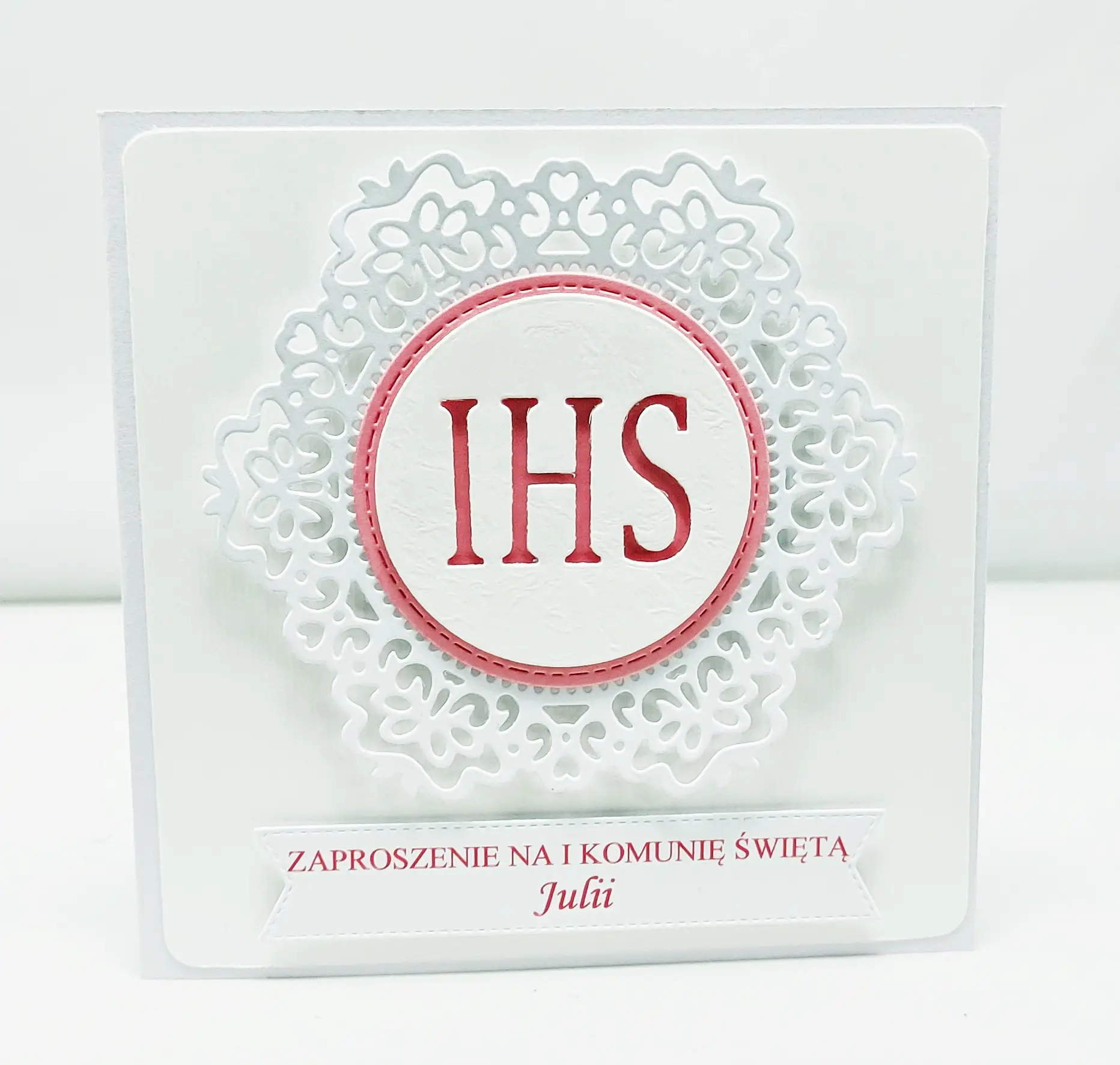 Różowe zaproszenie na Pierwszą Komunię Świętą z białym koronkowym ornamentem i symbolem IHS, kwadratowe, na papierze wizytówkowym, elegancka dziewczęca wersja zaproszenia.