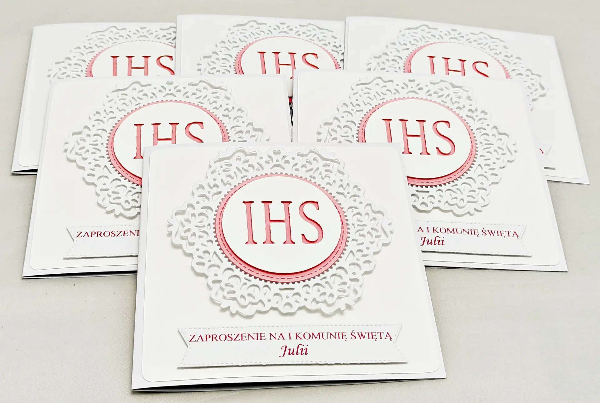 Kilka różowych zaproszeń na Pierwszą Komunię Świętą z koronkowym ornamentem i IHS, ułożonych obok siebie, widok z góry, elegancka dziewczęca kolorystyka.