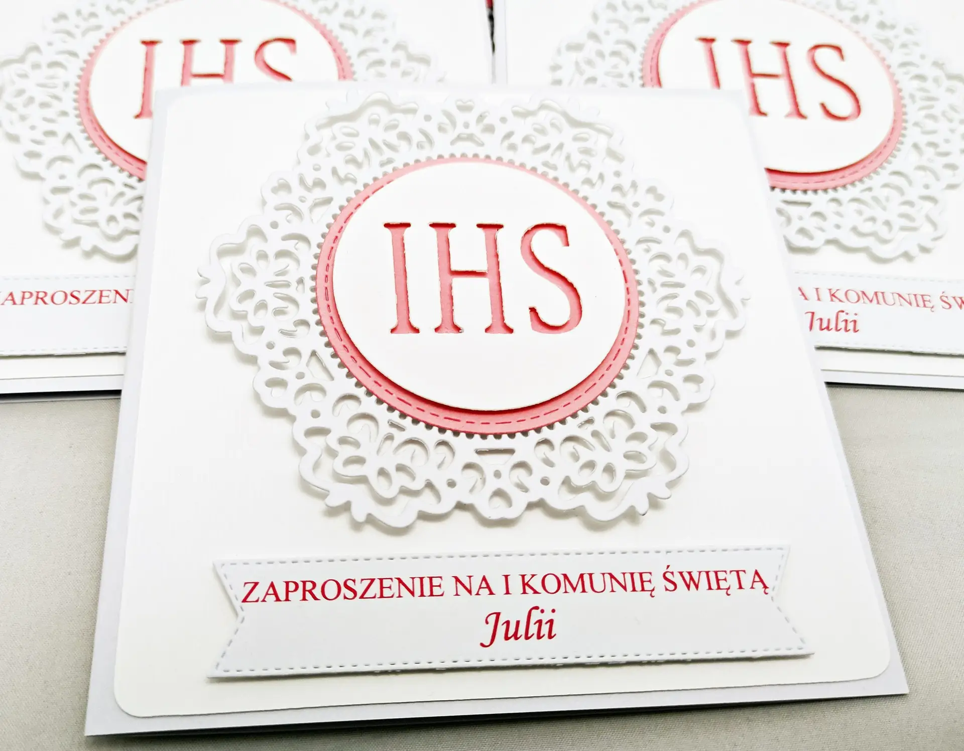 Zbliżenie różowego zaproszenia komunijnego z białym koronkowym ornamentem i symbolem IHS, widoczne detale ornamentu.