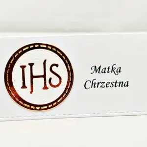 Winietka komunijna z motywem IHS, wykonana z białego papieru w połączeniu z lustrzanym złotem, ustawiona pojedynczo na stole