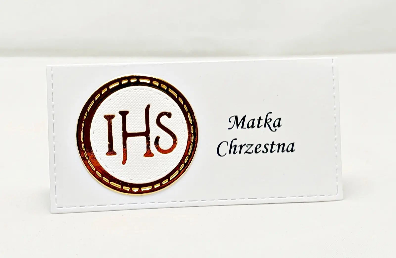 Winietka komunijna z motywem IHS, wykonana z białego papieru w połączeniu z lustrzanym złotem, ustawiona pojedynczo na stole