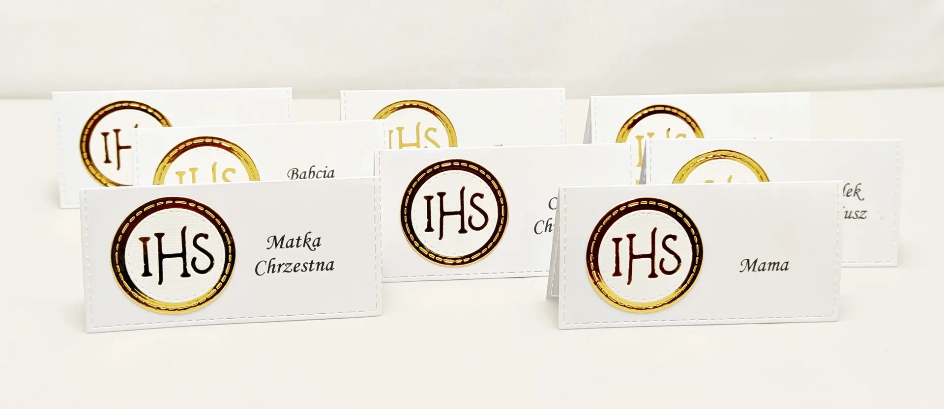 Kilka winietek komunijnych z motywem IHS, z białego papieru i lustrzanego złota, ustawionych w rzędzie, widok od przodu