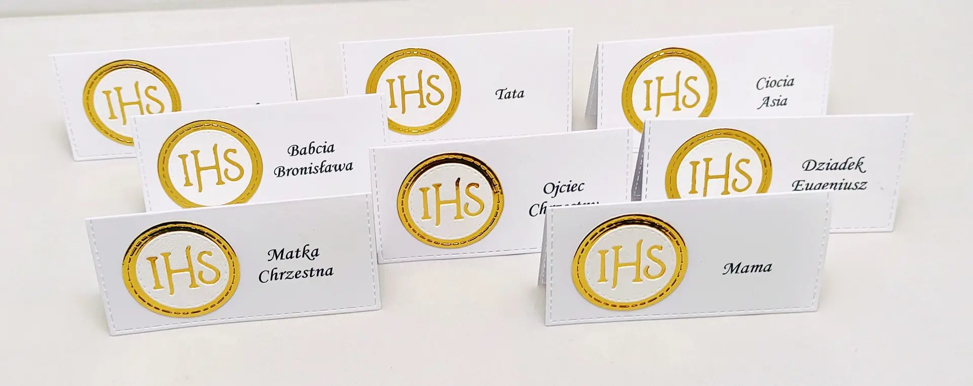 Winietki komunijne z motywem IHS z białego papieru i lustrzanego złota, uchwycone z góry, ułożone w rzędzie jako dekoracja stołu na komunii