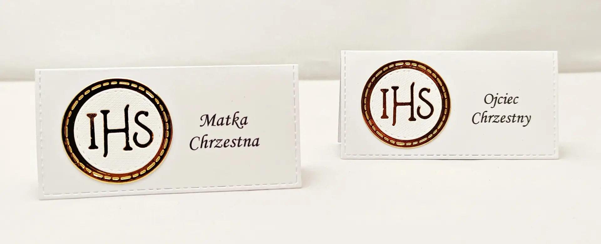 Dwie winietki komunijne z motywem IHS z białego papieru i lustrzanego złota, ustawione obok siebie jako dekoracja stołu na przyjęciu komunijnym