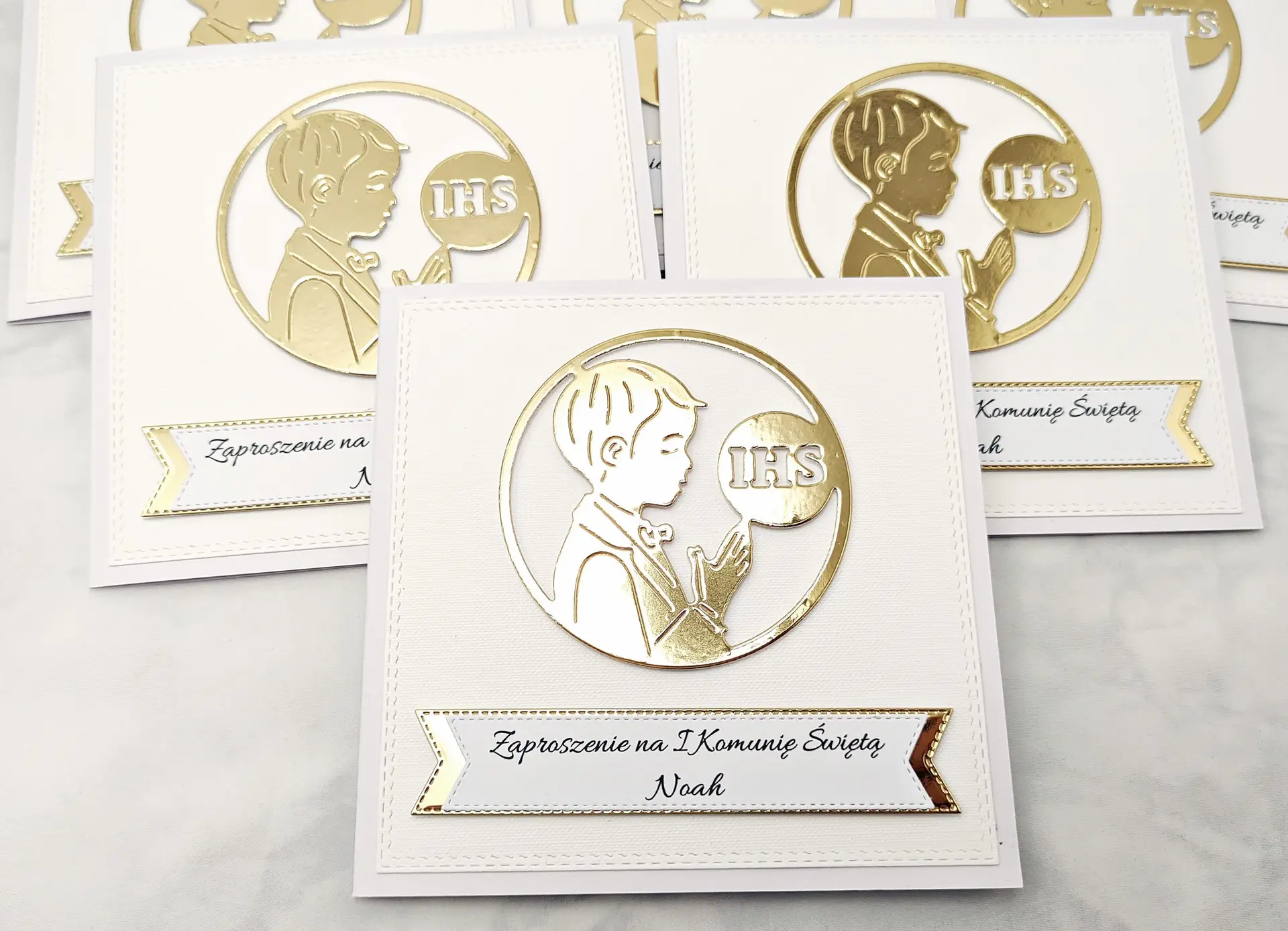 Trzy biało-złote zaproszenia komunijne dla chłopca z sylwetką chłopca i IHS, bez złotego tła, sfotografowane z góry w prostej kompozycji.