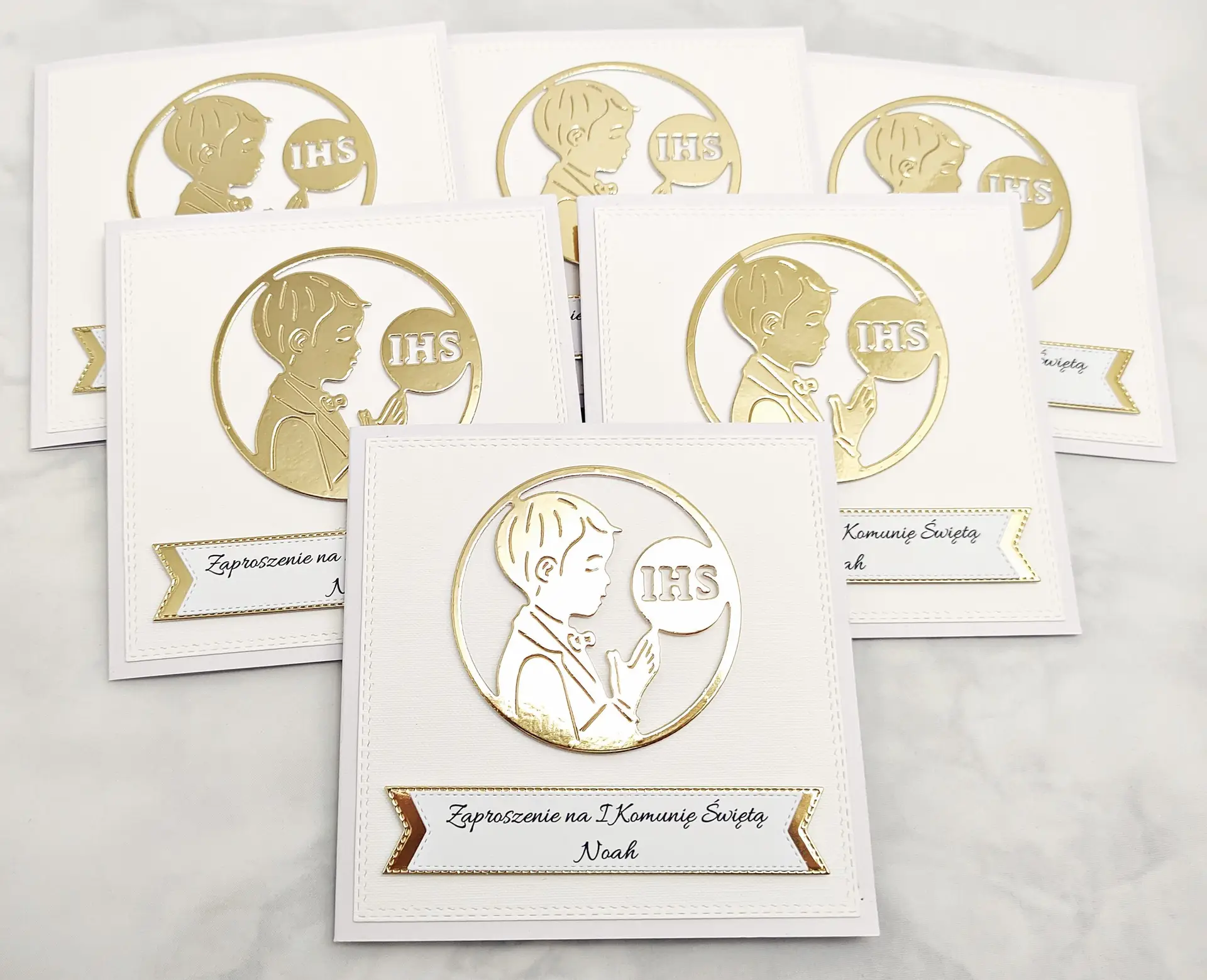 Kilka zaproszeń na Pierwszą Komunię Świętą dla chłopca z sylwetką chłopca i symbolem IHS, bez złotego tła, ułożonych obok siebie, widok z góry w zbliżeniu.