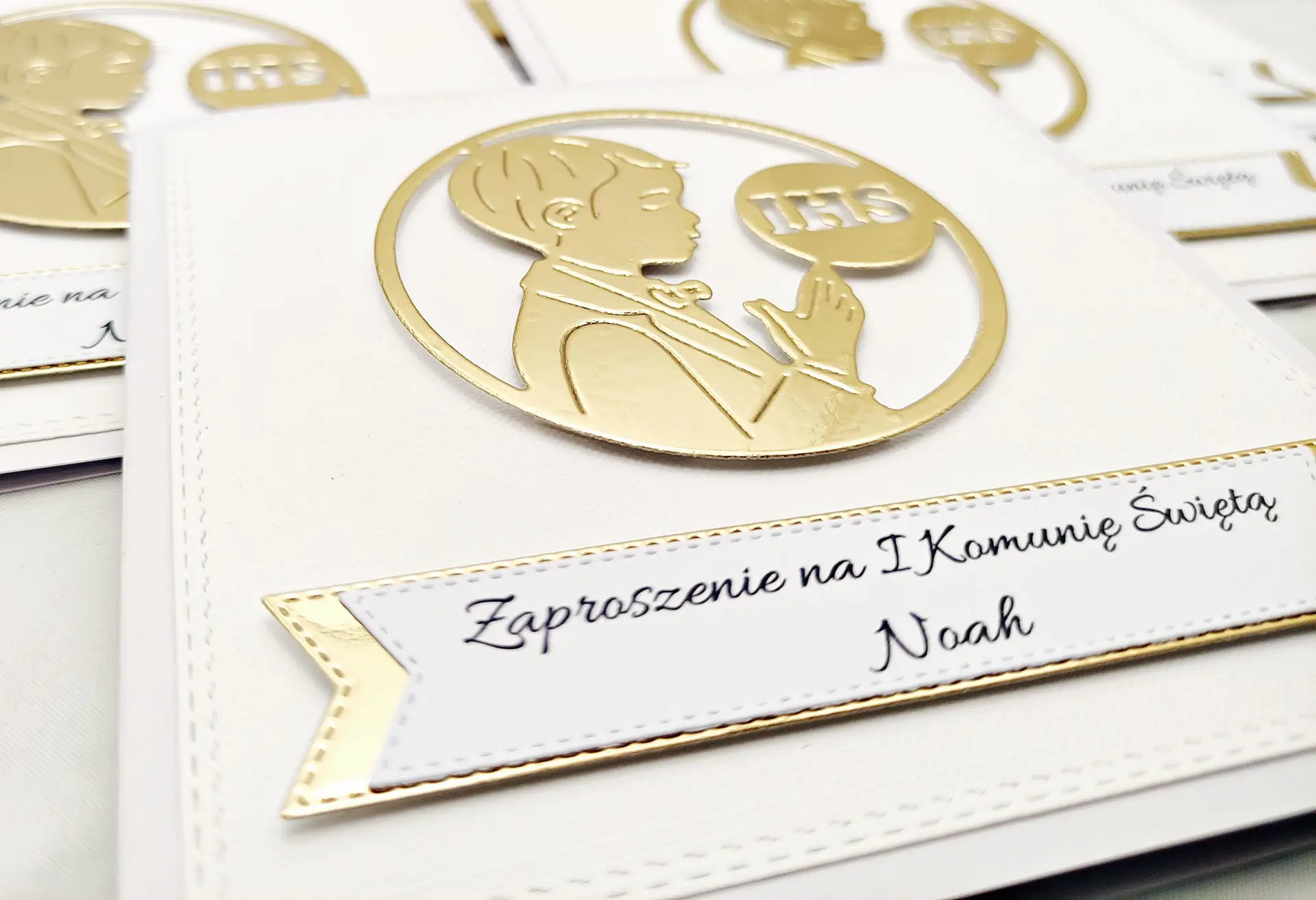 Zbliżenie zaproszenia komunijnego dla chłopca z sylwetką modlącego się chłopca i symbolem IHS na białym tle, bez lustrzanej złotej ramki.