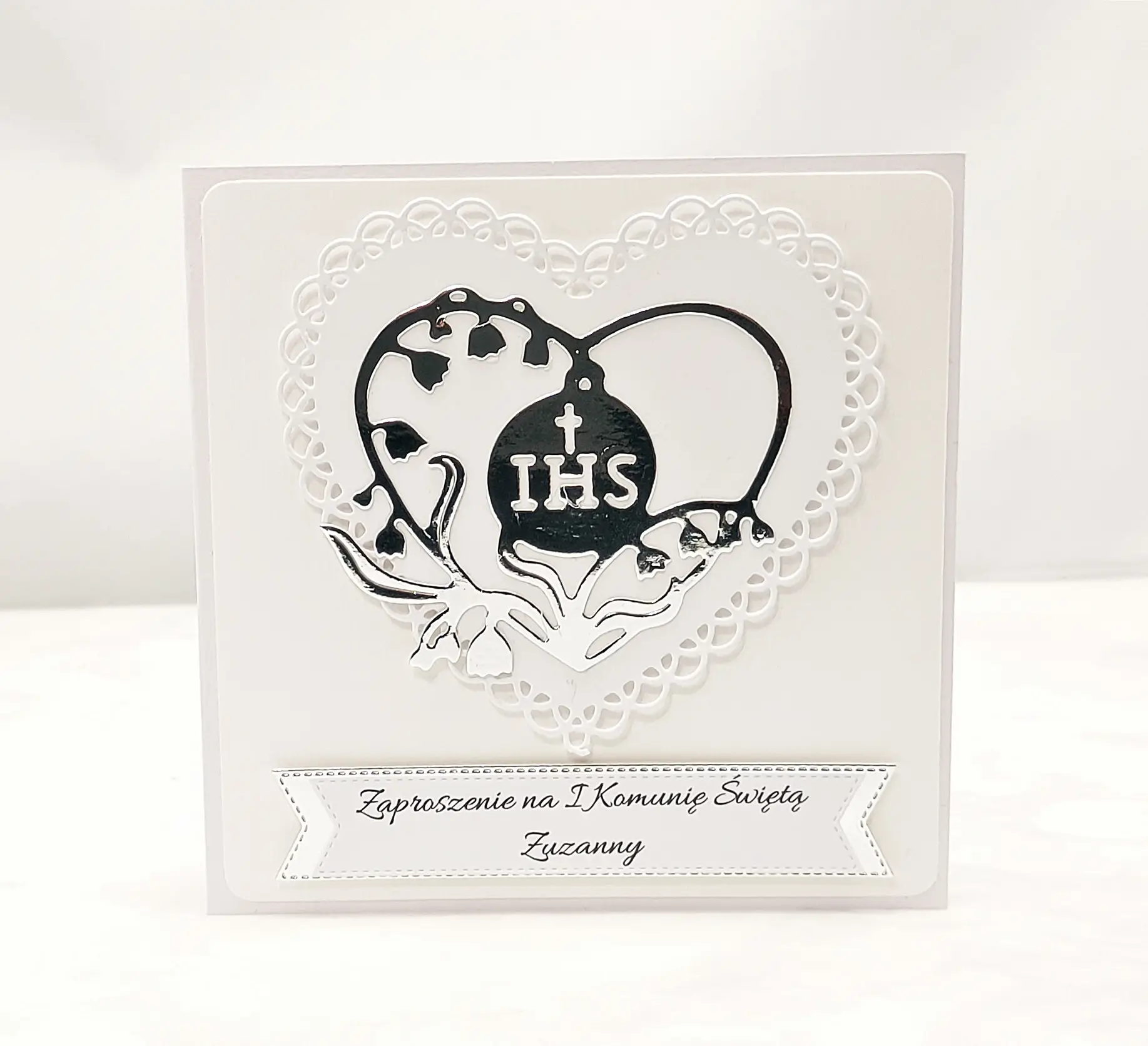 Biało-srebrne zaproszenie na Pierwszą Komunię Świętą z ażurowym sercem i sercem z konwalii wokół symbolu IHS, pojedyncze zaproszenie sfotografowane od frontu.