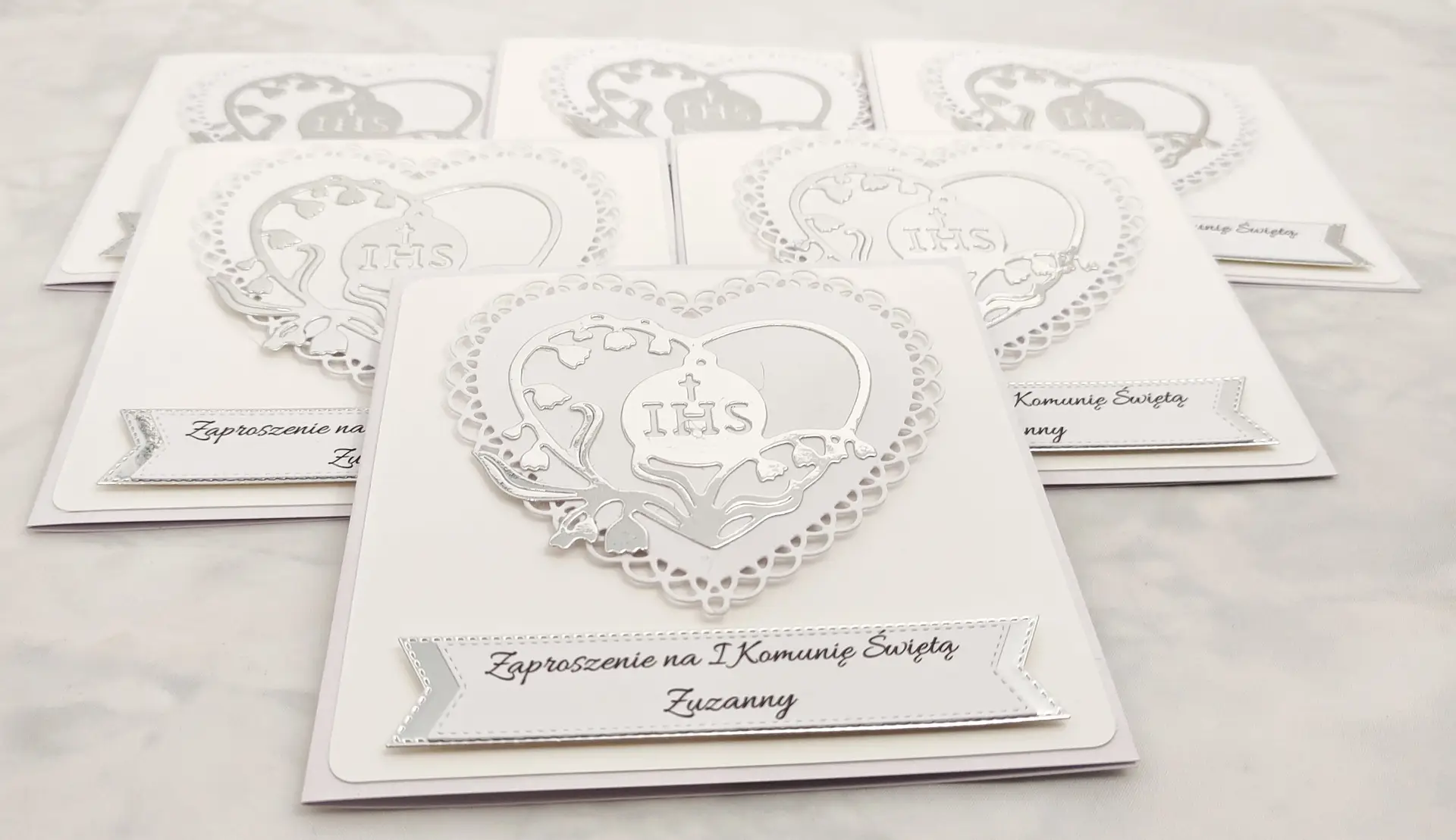 Kilka biało-srebrnych zaproszeń na Pierwszą Komunię Świętą z ażurowym sercem i sercem z konwalii z symbolem IHS, sfotografowanych z góry.
