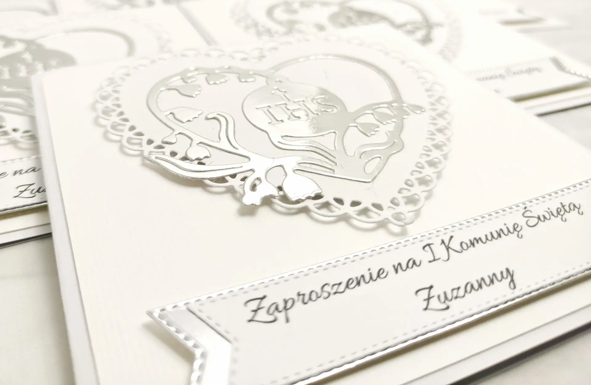 Zbliżenie biało-srebrnego zaproszenia komunijnego z ażurowym sercem i wewnętrznym sercem ułożonym z konwalii z symbolem IHS, widoczne subtelne detale wycinania.