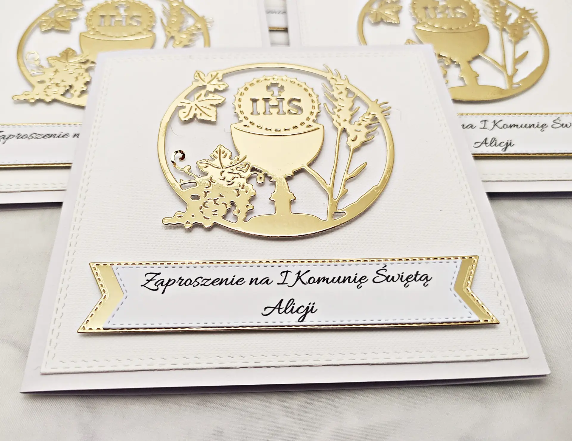 Zbliżenie zaproszenia komunijnego z kielichem i symbolem IHS w okręgu, na białym tle, widoczne delikatne złote akcenty i dekoracyjna kompozycja.